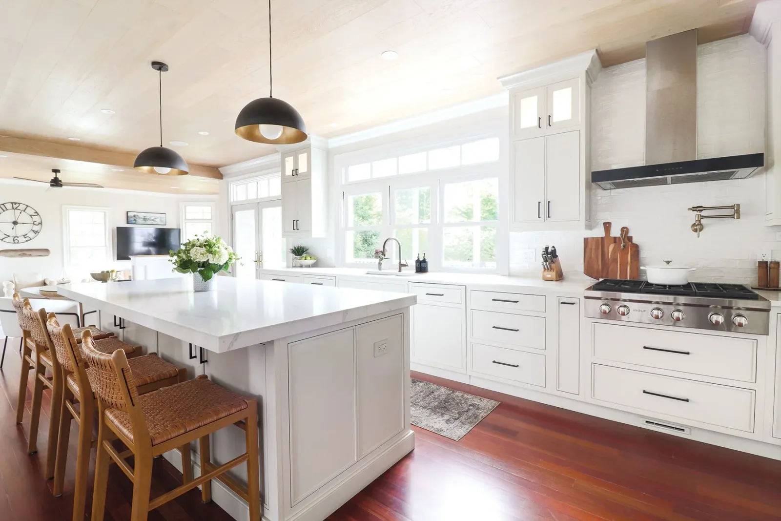 Modern kitchen with white cabinets, a large island with a white marble countertop, wooden bar stools, and stainless steel appliances. Bright natural light from large windows, hanging black and gold pendant lights, and hardwood floors.