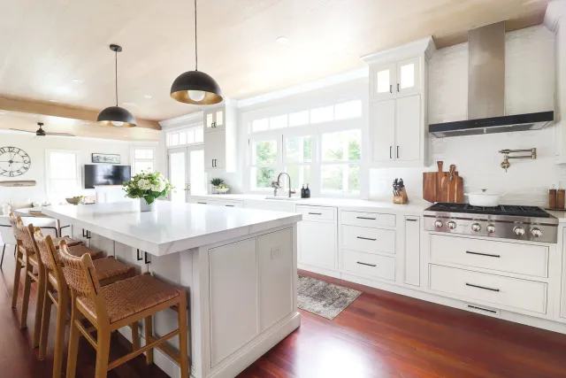 Modern kitchen with white cabinets, a large island with a white marble countertop, wooden bar stools, and stainless steel appliances. Bright natural light from large windows, hanging black and gold pendant lights, and hardwood floors.