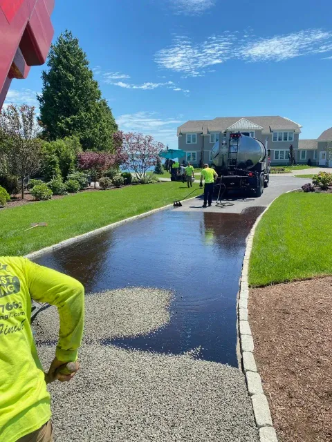 Asphalt sealing driveway with workers in bright vests and a tanker truck on a sunny day.