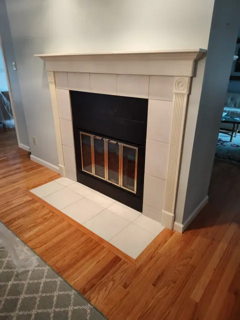 A white wooden fireplace mantle with a black insert and light-colored square tile surround, set on hardwood floors.