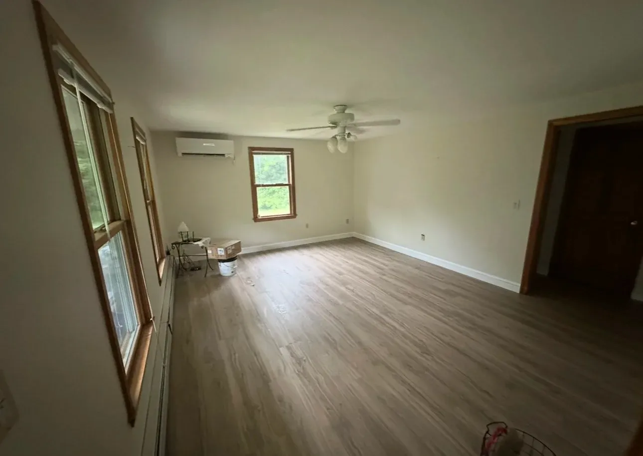 Empty living room with three windows, ceiling fan, air conditioner, hardwood floor, small side table with lamp near the windows, and doorway leading to another room.