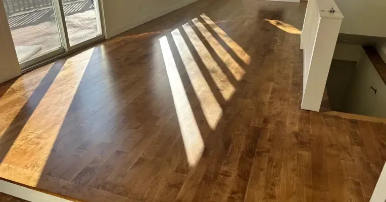 Sunlit empty hardwood room with long window shadows and a stair opening on the right