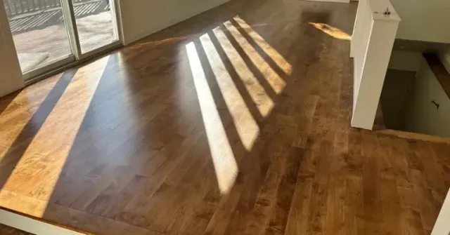 Sunlit empty hardwood room with long window shadows and a stair opening on the right