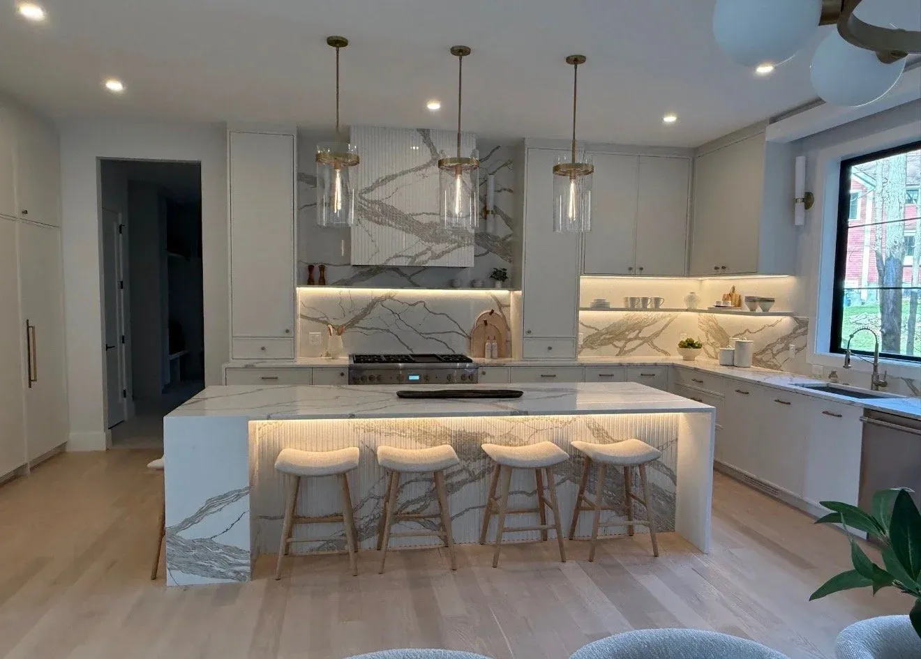 Modern kitchen with marble countertops, white cabinets, a large island with seating, three pendant lights above, and a window overlooking trees.