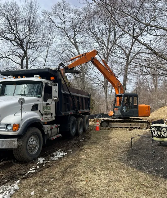 An orange excavator loads dirt into the back of a white dump truck parked on a dirt path near bare trees.
