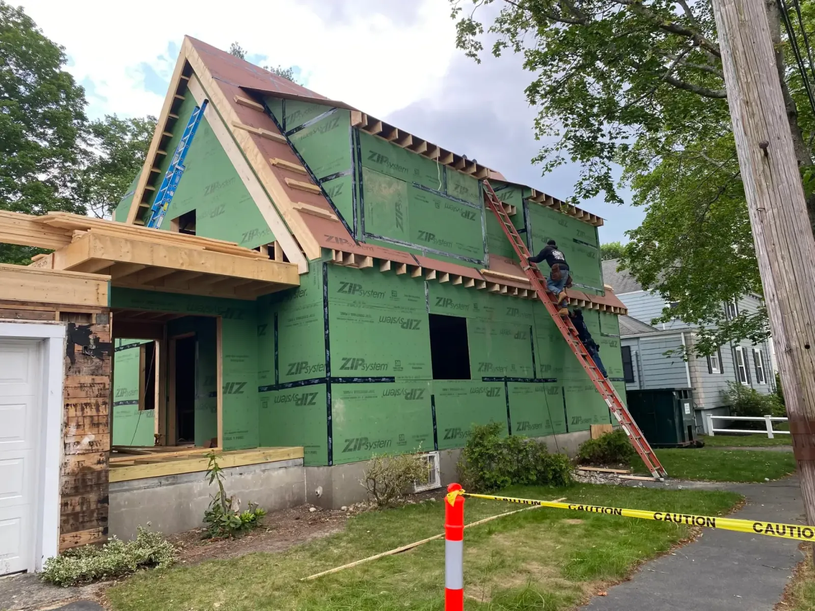 Construction workers applying ZIP sheathing to a custom home in Wakefield, MA, featuring a wooden ladder and partially completed exterior with green sheathing.