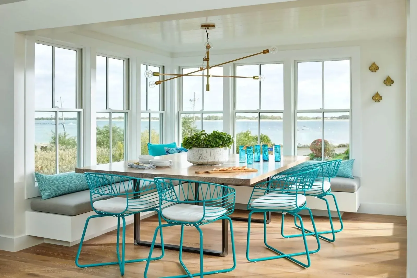 Bright dining nook with large windows overlooking water, featuring a wooden table, turquoise wireframe chairs with white cushions, built-in window seat with cushions, a modern gold chandelier, and decorative blue glassware.