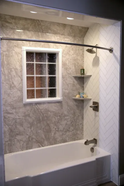 A white bathtub with silver fixtures, built-in shelves, white and gray walls, and a window.