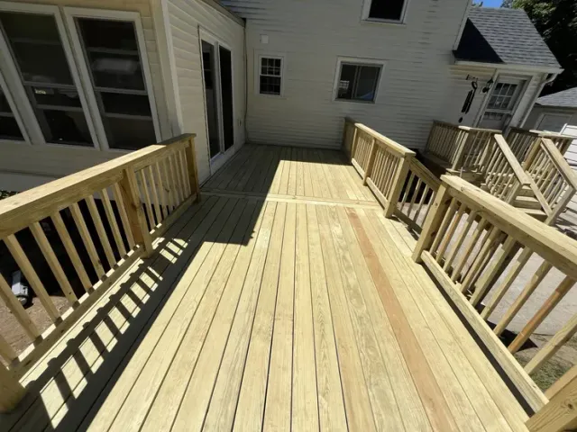 Wooden rooftop deck with railings beside a house, lit by bright sunlight
