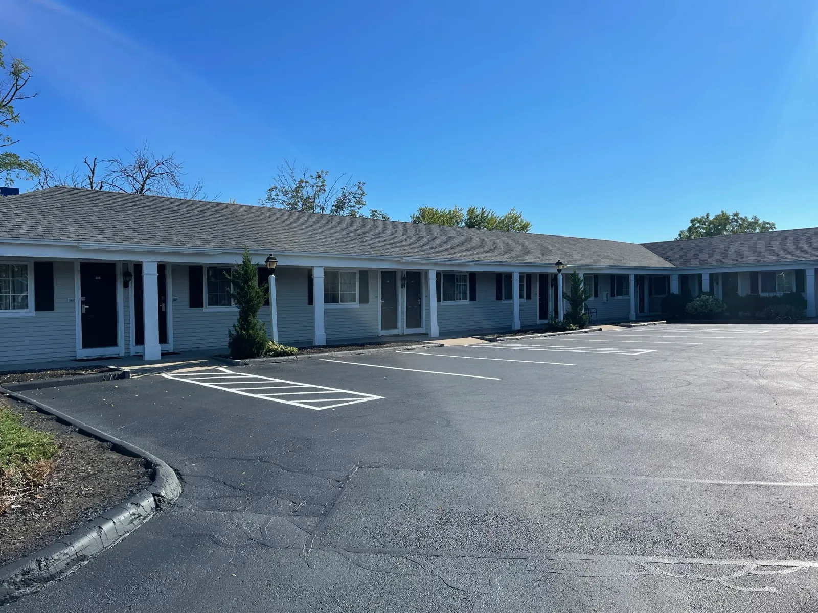Exterior view of a single-story apartment complex with a parking lot. The building has gray siding, black shutters, and white columns, surrounded by a few small trees. The parking lot has several unoccupied spaces and is under a clear blue sky.