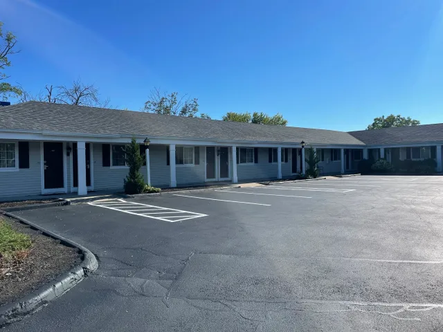 Exterior view of a single-story apartment complex with a parking lot. The building has gray siding, black shutters, and white columns, surrounded by a few small trees. The parking lot has several unoccupied spaces and is under a clear blue sky.