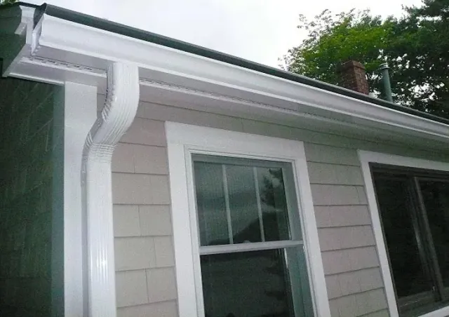 White aluminum gutters and a downspout attached to the corner of a house with light gray shingled siding.