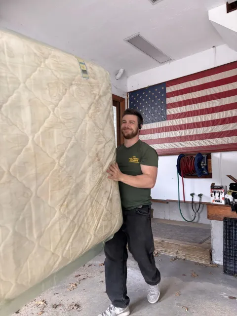 Person carrying a mattress in a garage with an American flag on the wall.