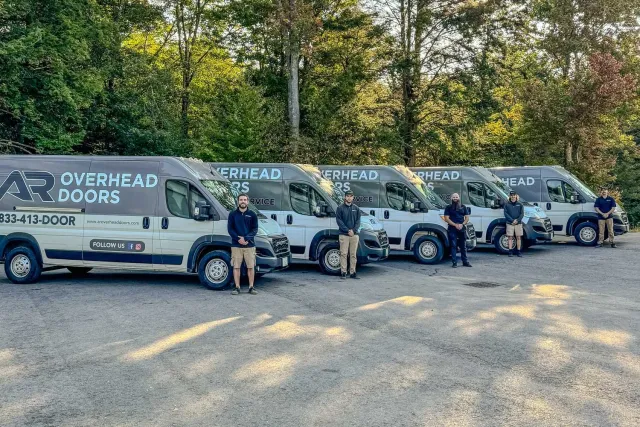 Five men standing in front of AR Overhead Doors service vans parked in a wooded area.