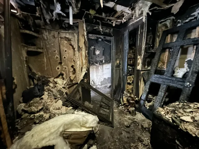 A burnt, damaged interior room with charred walls, ceiling, and furnishings, showing extensive fire damage.