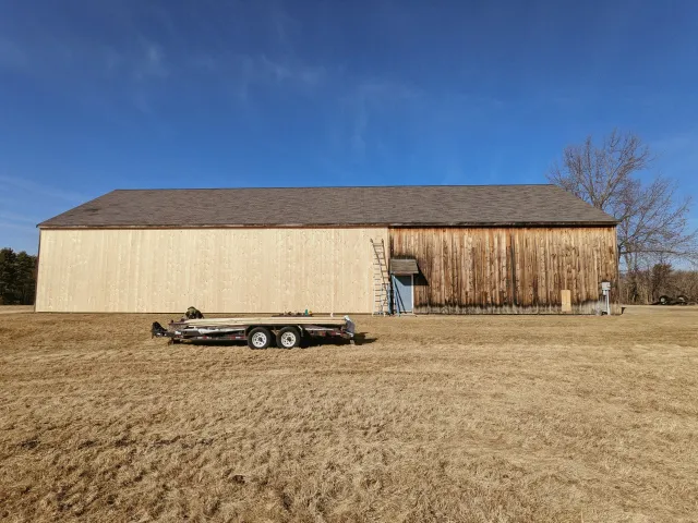 A large wooden barn under a clear blue sky with a leafless tree nearby. A trailer with construction materials is parked in front of the barn.