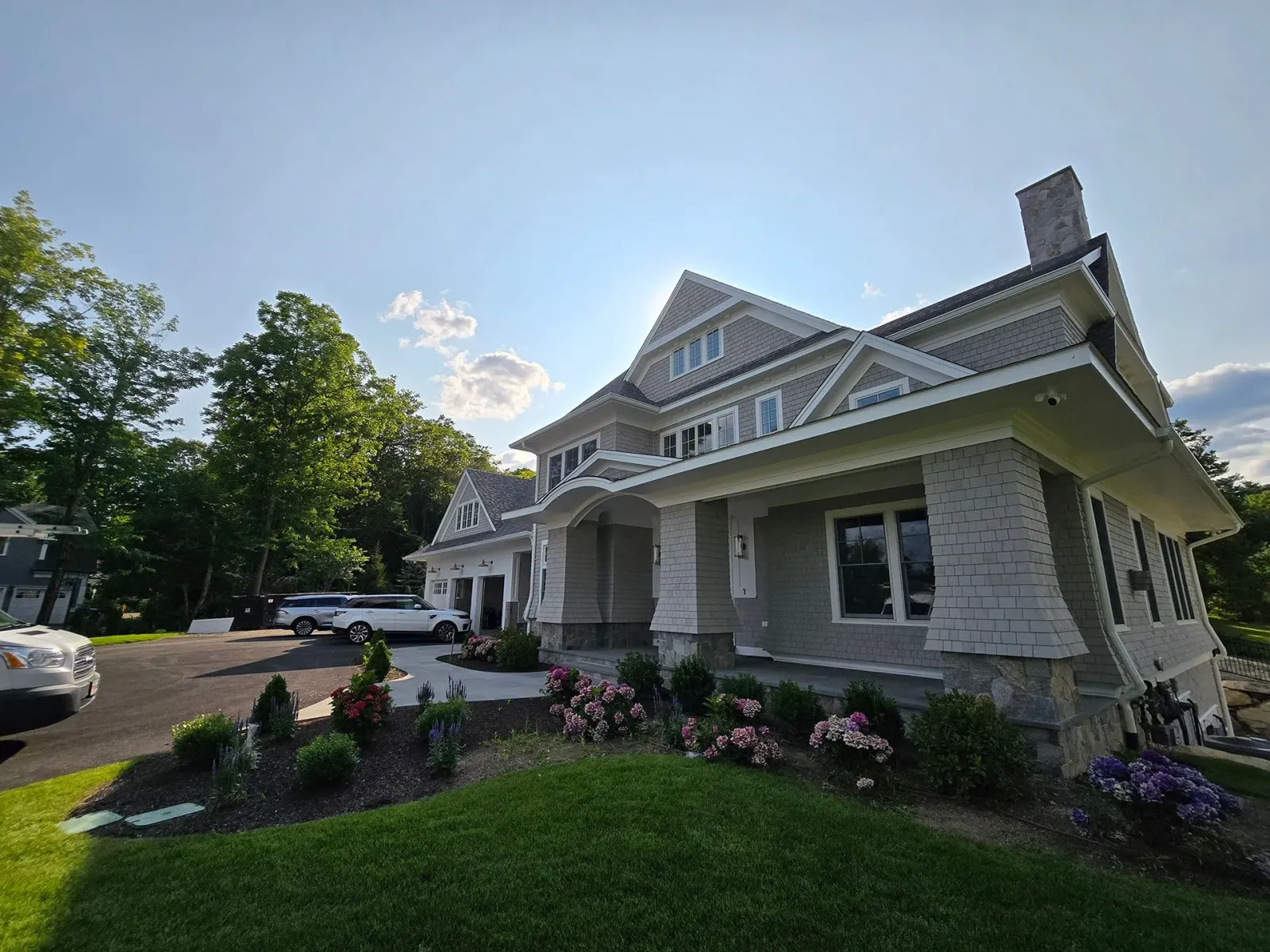Historic white house with porch, flower beds, and parked cars on a sunny day