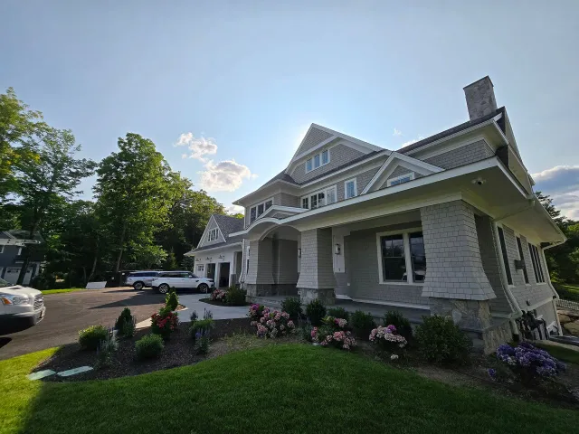 Historic white house with porch, flower beds, and parked cars on a sunny day