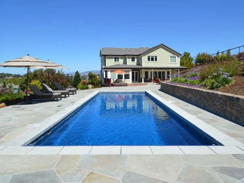 A backyard with a swimming pool, lounge chairs with a large umbrella, and a two-story house with a porch, surrounded by landscaped gardens and clear blue sky.
