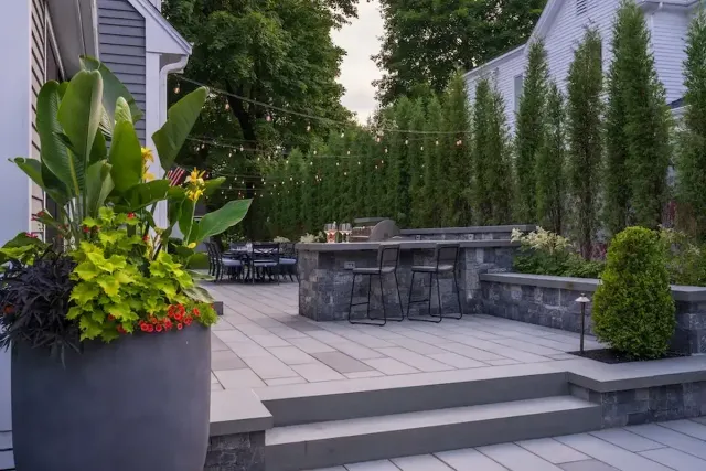 Backyard patio with a gray stone bar, string lights overhead, and potted plants along the edge.