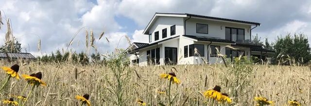A modern white two-story hilltop home with large windows sits behind a field of tall grasses and yellow wildflowers under a partly cloudy sky. Trees are visible in the background.
