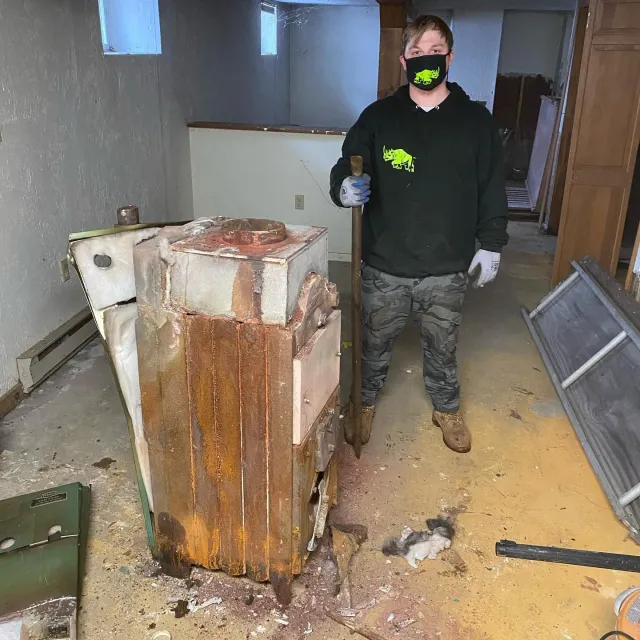 Man standing near a damaged heater in a basement, holding a tool. He wears a mask and work clothes.