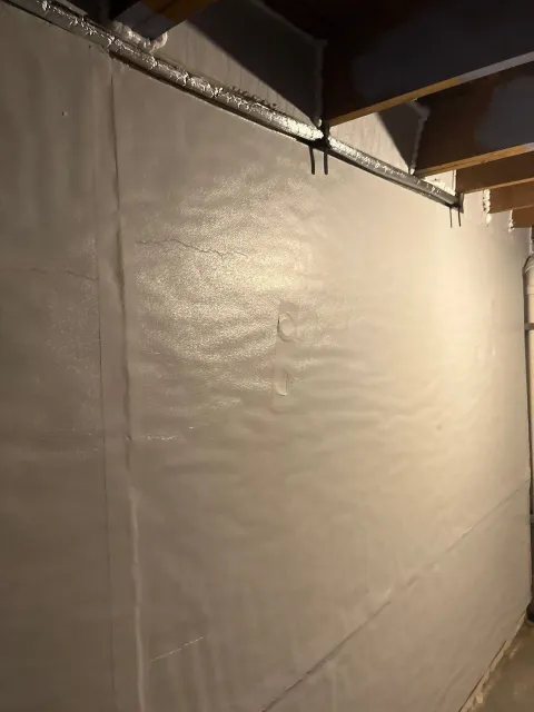 Dimly lit unfinished wall with beige insulation or tarp beneath exposed ductwork and ceiling beams