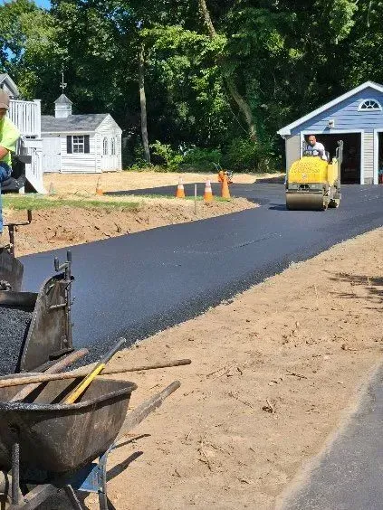 A person operates a yellow steamroller to pave a new asphalt driveway in front of a residential house.