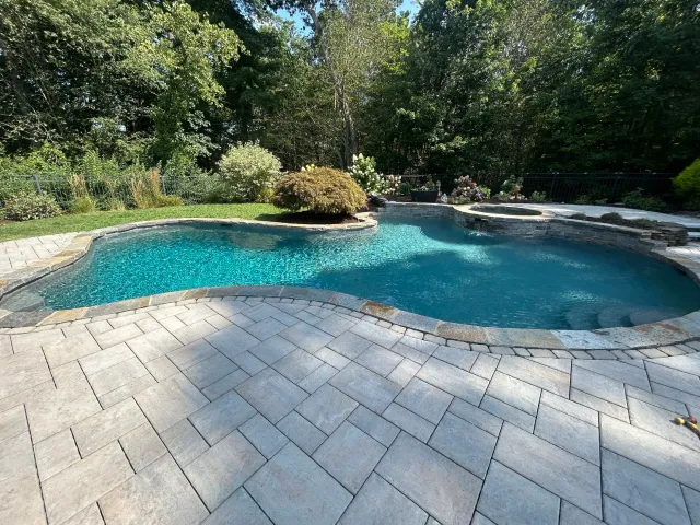 A backyard with a kidney-shaped swimming pool surrounded by a stone patio, lush trees, and flower bushes.