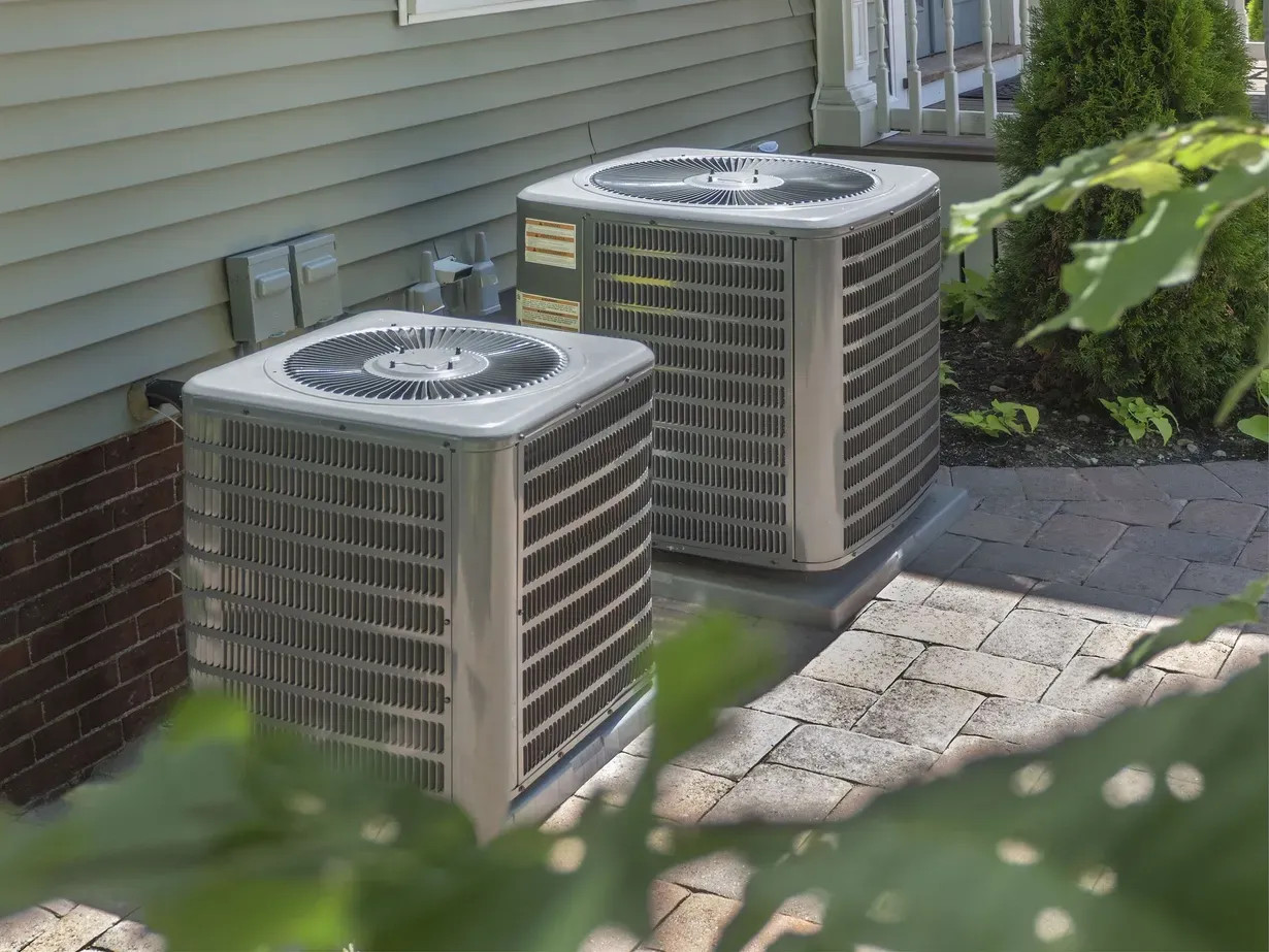 Two outdoor air conditioning units beside a house wall, partially framed by leaves and a brick patio.