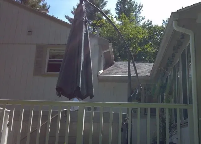 A closed dark patio umbrella standing on a wooden deck next to the side of a house.