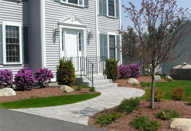 Suburban house entrance with grey siding, flowering bushes, and a landscaped path. Neat shrubs and small trees enhance the well-maintained front yard.