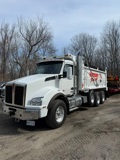 A white Kenworth dump truck parked on a dirt lot, featuring a large chrome bumper and an attached red trailer.