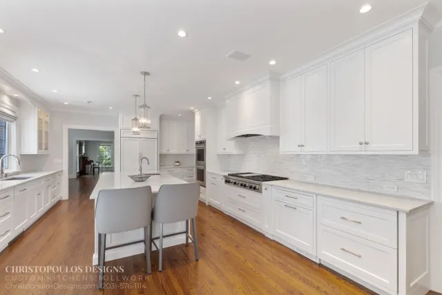 Bright white kitchen with island, white cabinets, stainless steel appliances, and hardwood floors, illuminated by recessed lighting and hanging pendant lights.