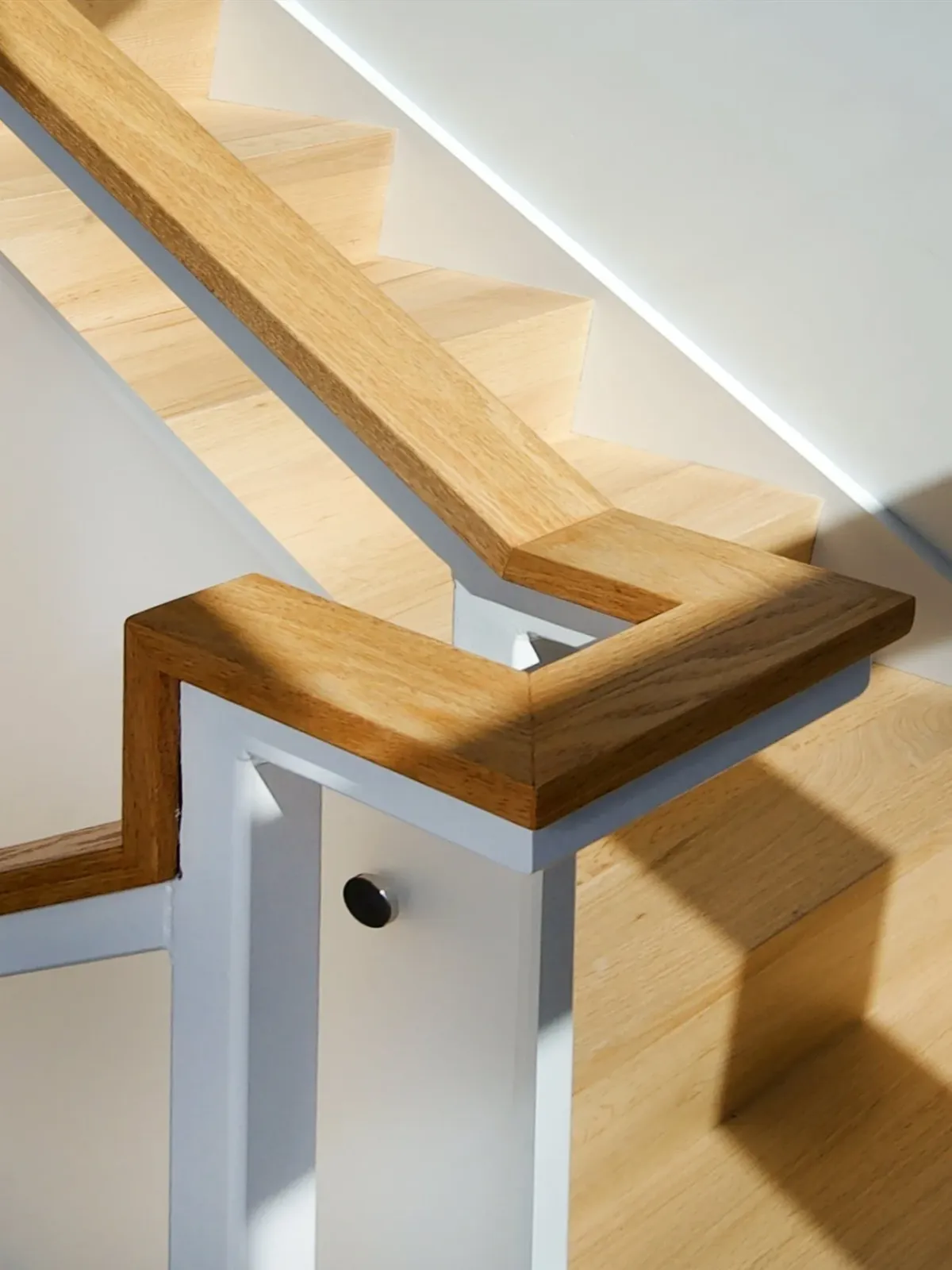 An angled view of a staircase railing with wooden handrails and white balusters, showing sunlight and shadows.