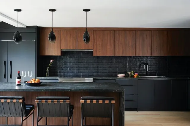 Modern kitchen with dark cabinetry, black subway tile backsplash, wood upper cabinets, and a black island with seating, decorated with flowers, wine, and a fruit bowl under three pendant lights.