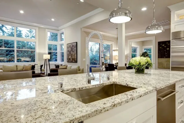 Granite countertop with stainless steel sink in a modern kitchen with large windows and pendant lights.