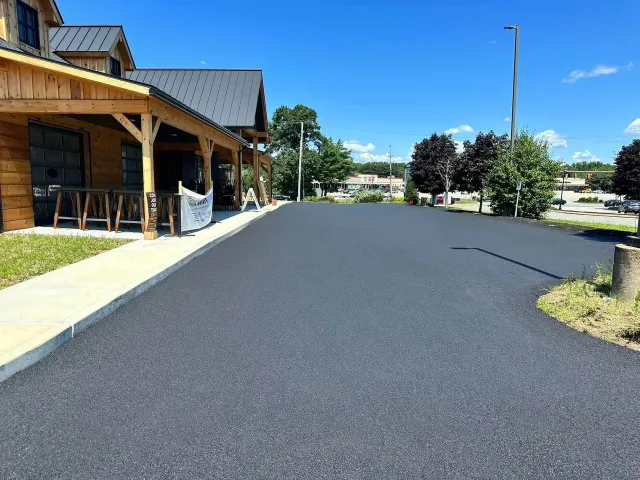 Newly paved asphalt parking lot in front of a wooden building with a porch and seating, under a blue sky.