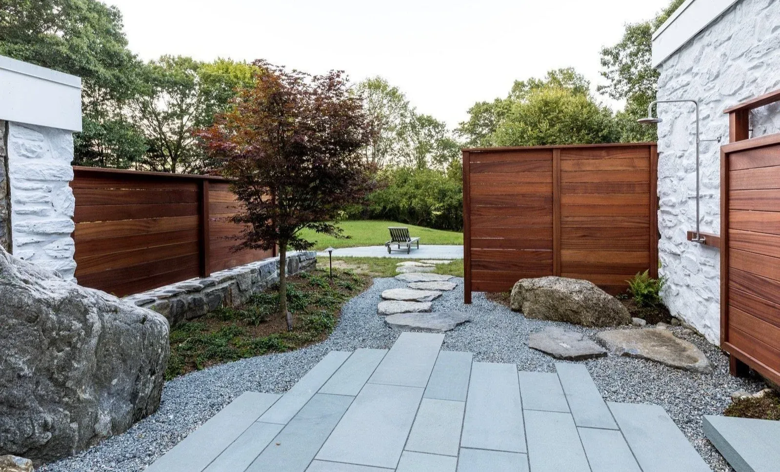 Backyard garden with a stone pathway, a small tree, and a bench in the distance, enclosed by wooden and white stone walls.