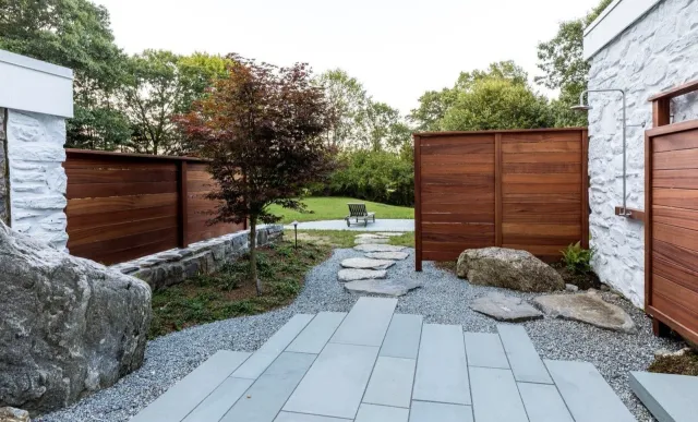 Backyard garden with a stone pathway, a small tree, and a bench in the distance, enclosed by wooden and white stone walls.