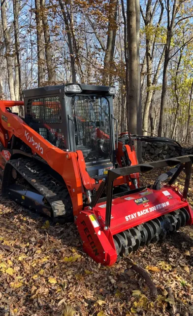 Orange Kubota track loader with a red mulcher attachment in a wooded area.
