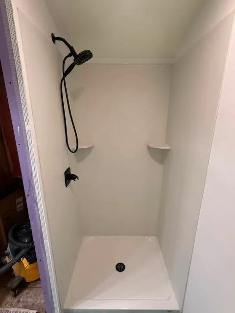Empty shower stall with white walls, showerhead, and built-in corner shelf.