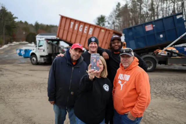 Group of five people taking a selfie outdoors with a large truck and an orange container in the background.