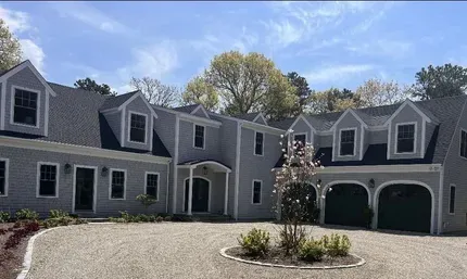 Large gray house with dormer windows, arched garage doors, and a gravel driveway under a clear sky