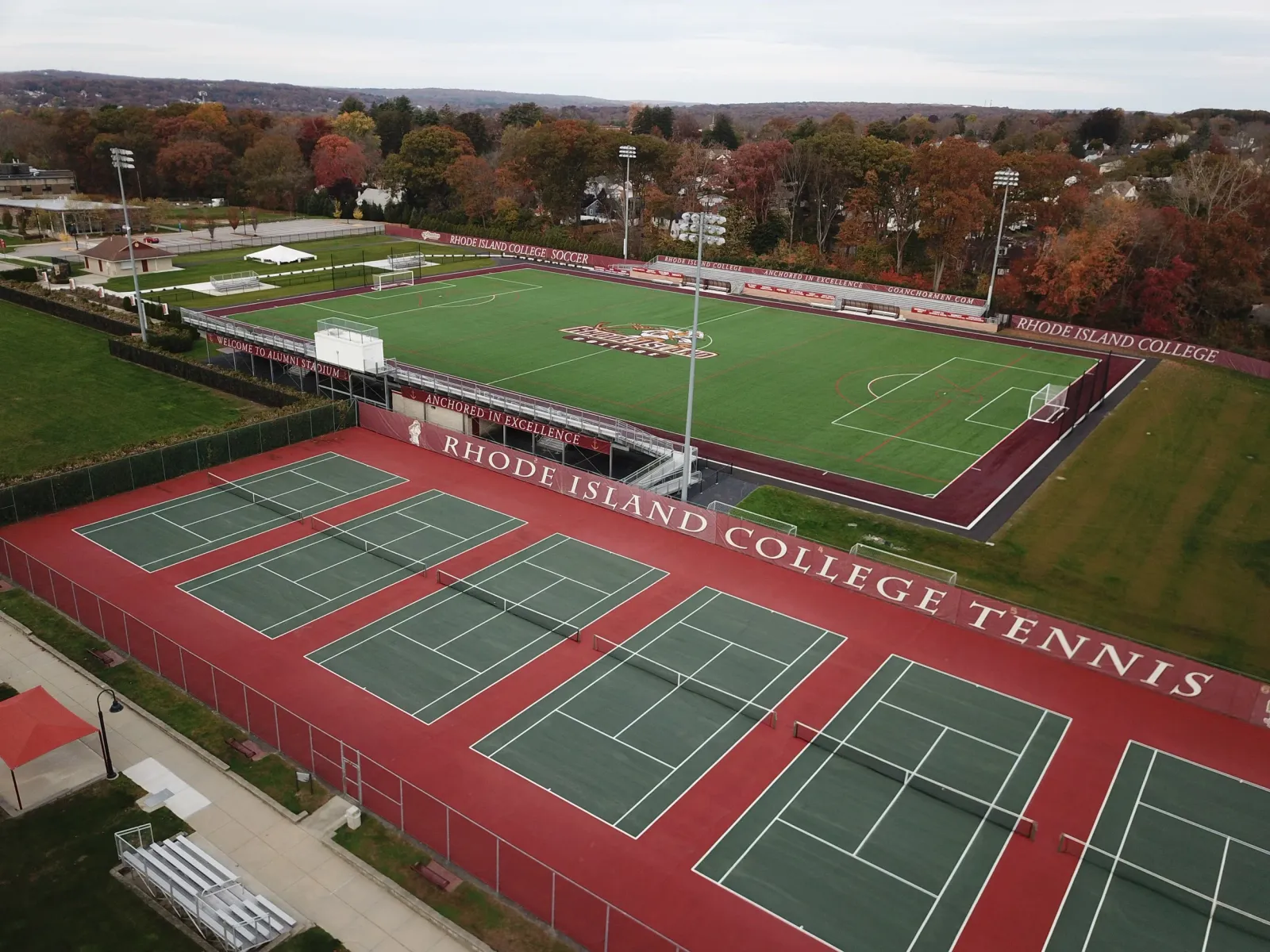 Aerial view of an athletic complex at Rhode Island College including a soccer field, tennis courts, and a running track, with fall foliage in the background.