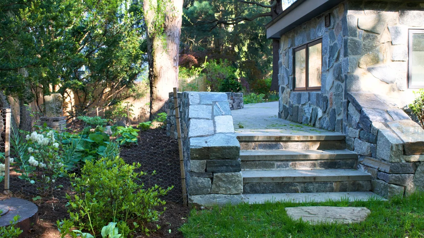 A stone building with large windows is surrounded by lush greenery. Stone steps lead to the entrance, with trees and shrubs in the background. A small garden is visible on the left, bordered by a wire mesh fence.