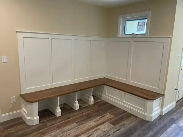 An L-shaped wooden bench with white paneling and a stained wood seat, situated against a wall in a room with wood floors.