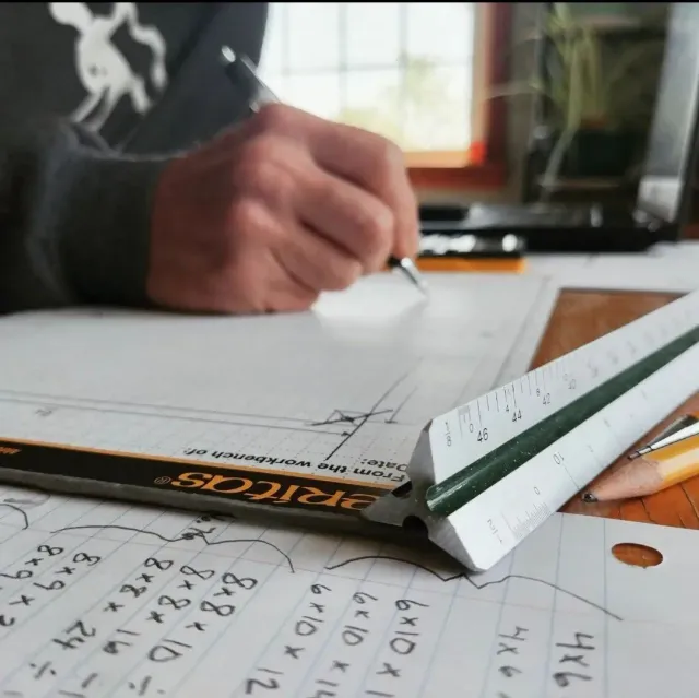 A person writing on graph paper with a pencil and a ruler, in a room with natural light from a window.