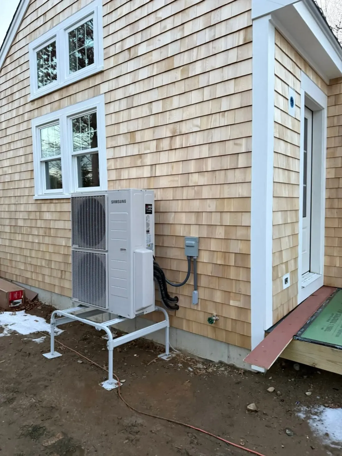 A mini-split heat pump unit stands on a white metal frame outside a house clad in light-colored wooden shingles.