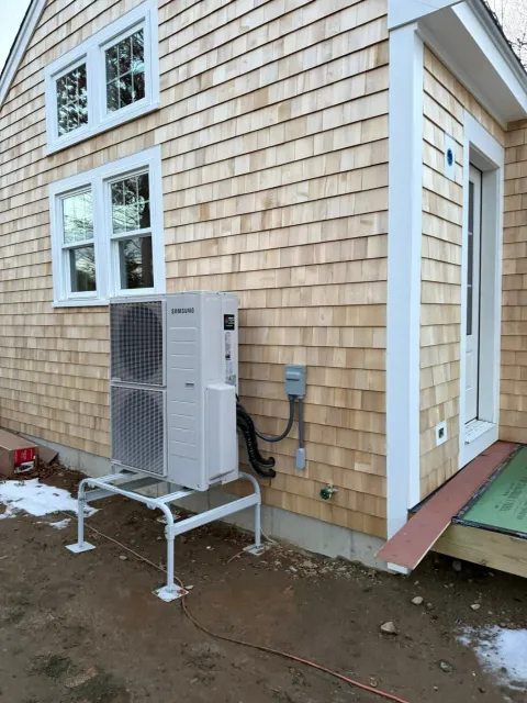 A mini-split heat pump unit stands on a white metal frame outside a house clad in light-colored wooden shingles.